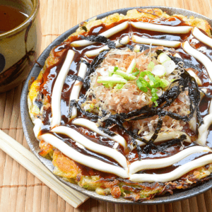 Okonomiyaki drizzled with mayonnaise and sauce, topped with bonito flakes and nori, beside a cup of tea