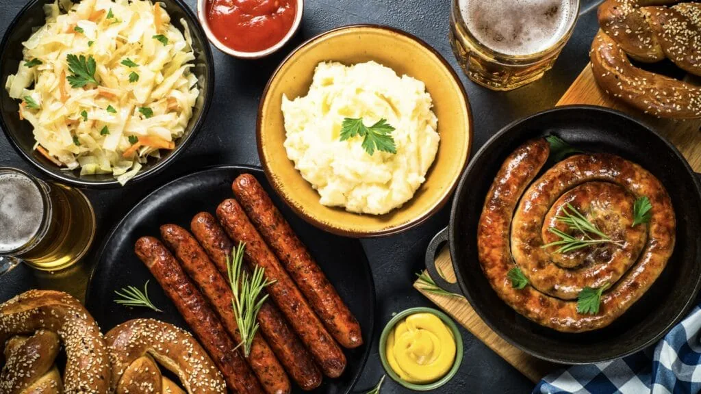 Overhead spread of bratwurst, a coiled sausage, mashed potatoes, coleslaw, pretzels and glasses of beer