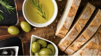 Bowl of olive oil with rosemary beside green olives and slices of crusty bread on a wooden board