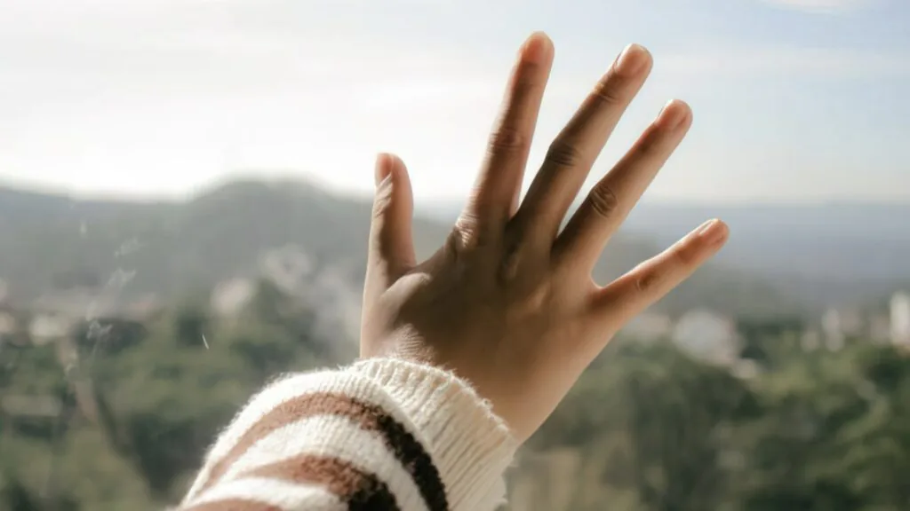 Open hand with five fingers spread, pressed against a window overlooking hills and sky