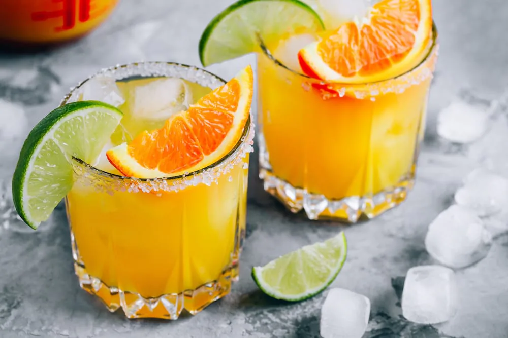 Two orange margaritas with salted rims, garnished with orange slices and lime wedges over ice
