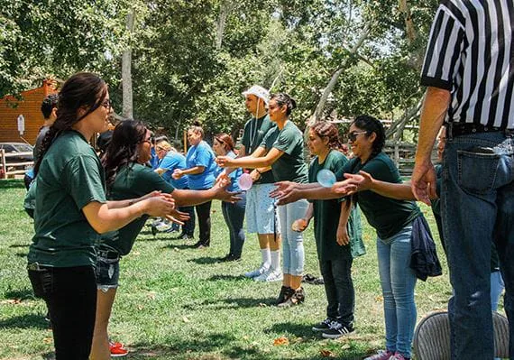 Two teams in green and blue shirts face off in an outdoor team-building game with a referee