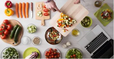 Overhead view of hands sliding chopped vegetables from a cutting board into a bowl beside a laptop