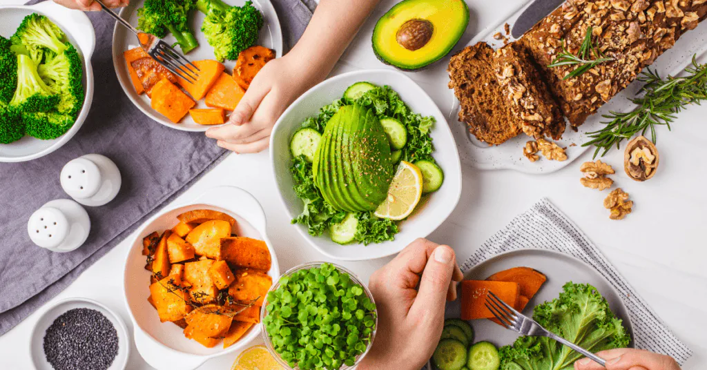 Overhead view of hands sharing bowls of avocado salad, roast sweet potato, broccoli and seeded bread