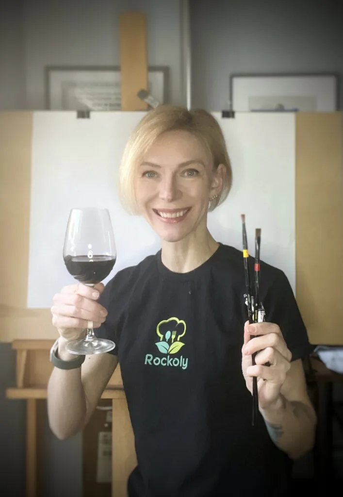 Paint and sip host in a Rockoly shirt holding a glass of red wine and paintbrushes beside an easel