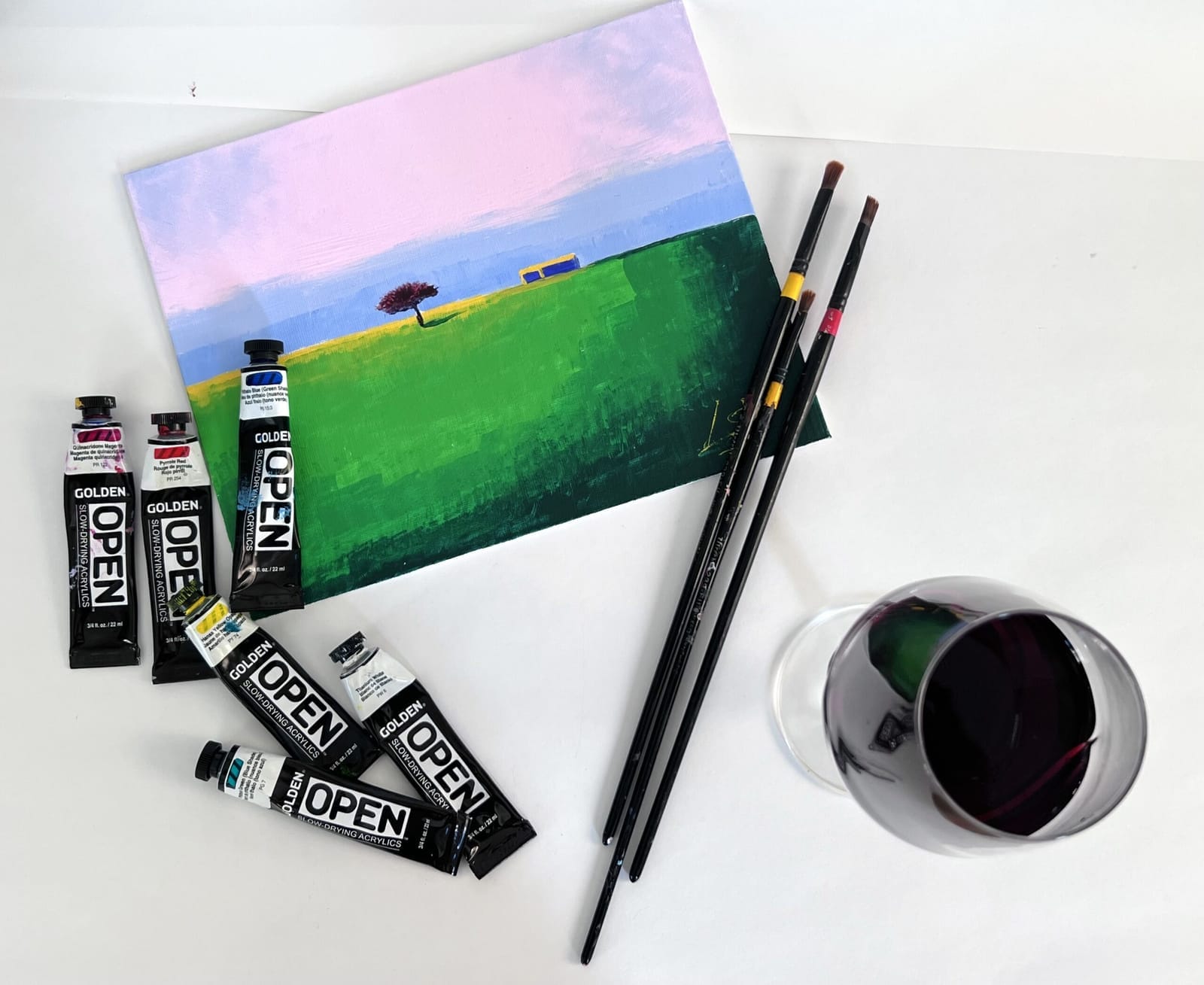 Finished landscape canvas with acrylic paint tubes, brushes and a glass of red wine