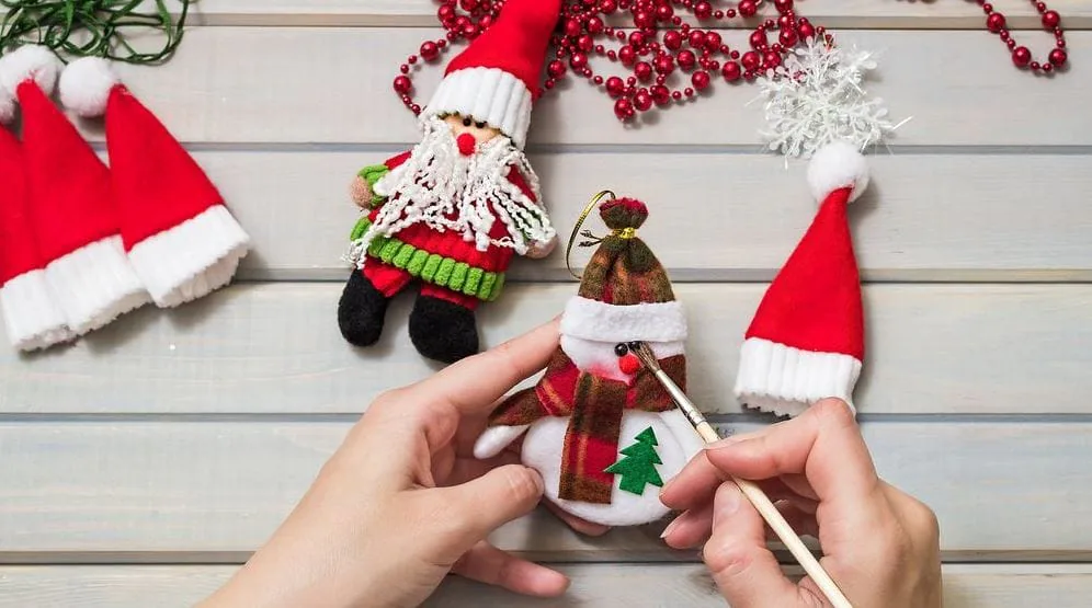 Hands painting a small snowman ornament with a brush among Santa hats and holiday beads
