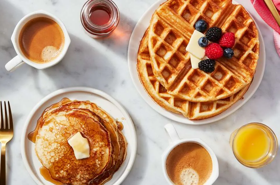 Overhead of a stack of buttered pancakes beside a waffle with berries, coffee and orange juice