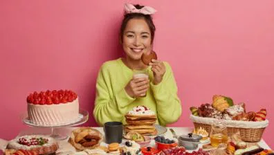 Smiling person in a green sweater surrounded by cake, pancakes and pastries against a pink backdrop