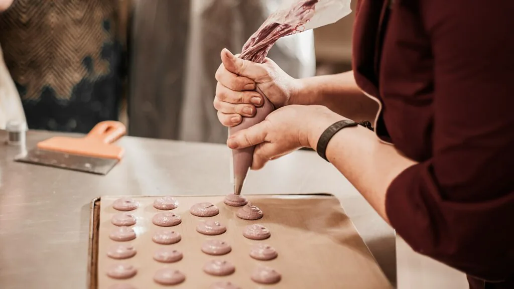 Hands piping rows of pink macaron shells onto a lined baking tray in a kitchen