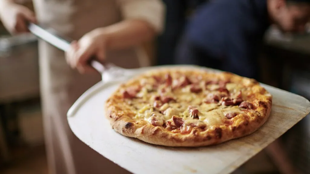 Freshly baked pizza topped with cheese and cured meat lifted on a wooden peel