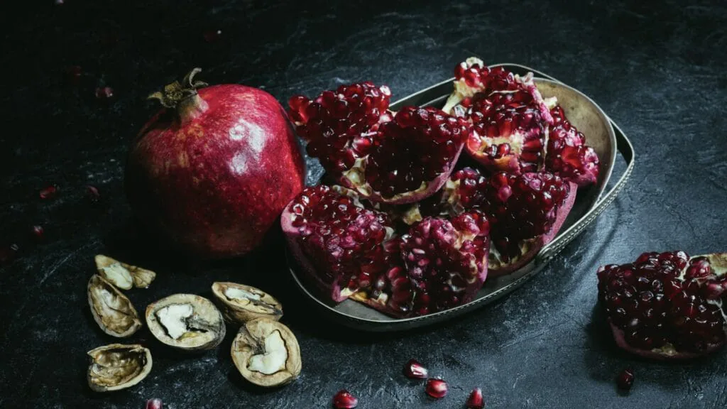 Whole and split pomegranates on a metal tray with cracked walnuts on a dark surface