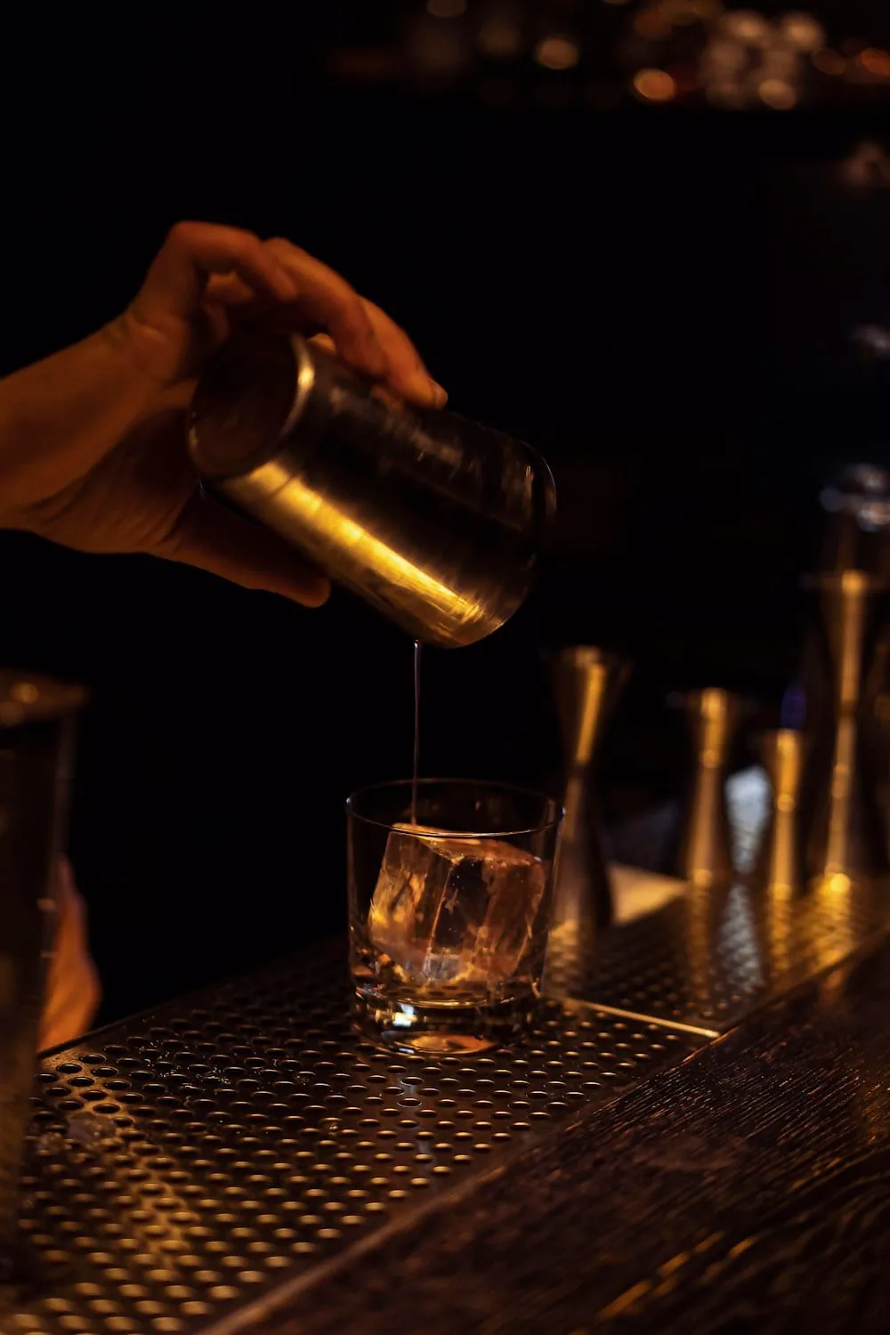 Pouring a cocktail from a shaker into an ice-filled rocks glass on a dimly lit bar