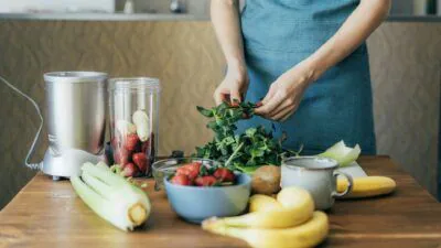 Person in an apron tearing fresh greens at a table of celery, bananas, strawberries and a blender