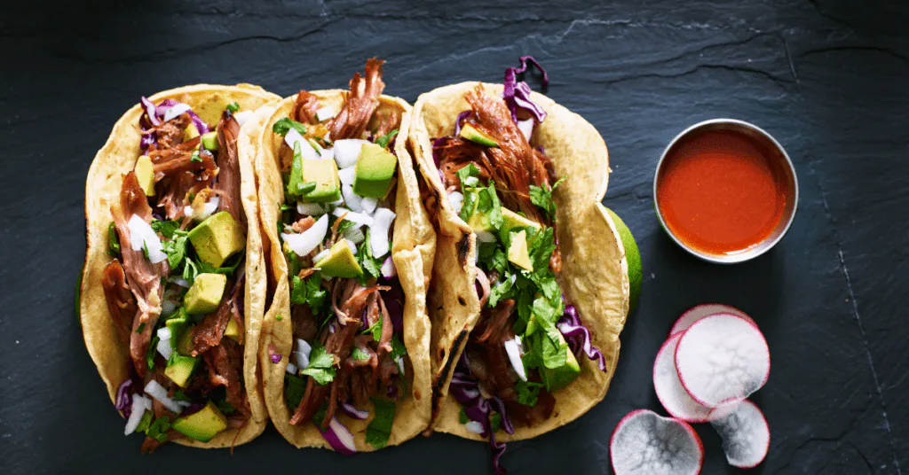 Three pulled pork tacos with avocado, red cabbage and cilantro beside a bowl of red salsa