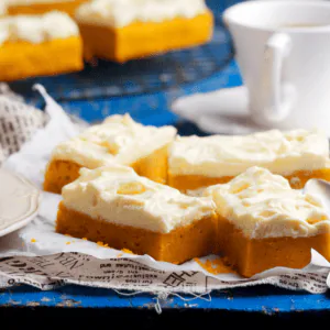 Squares of pumpkin bars topped with cream cheese frosting on a blue table beside a cup of coffee