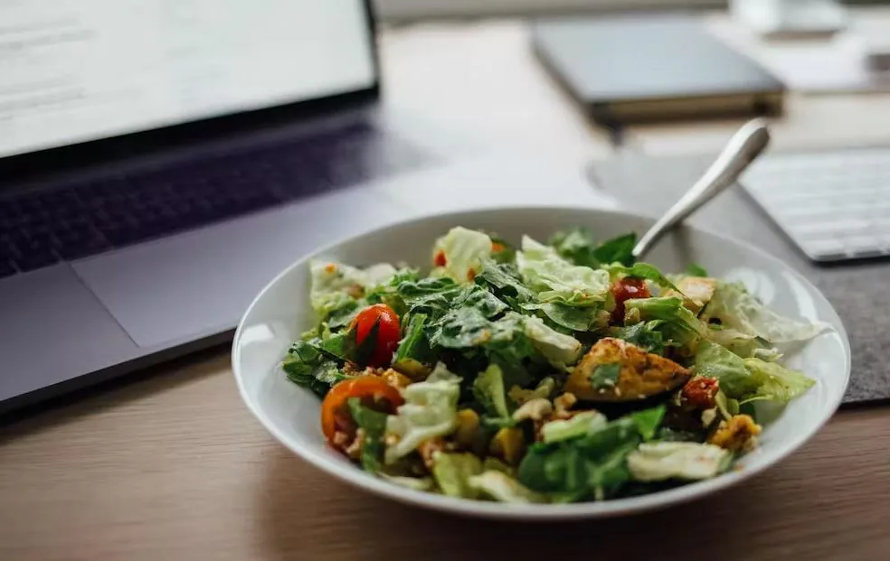 Bowl of green salad with tomatoes on a desk in front of an open laptop and notebook