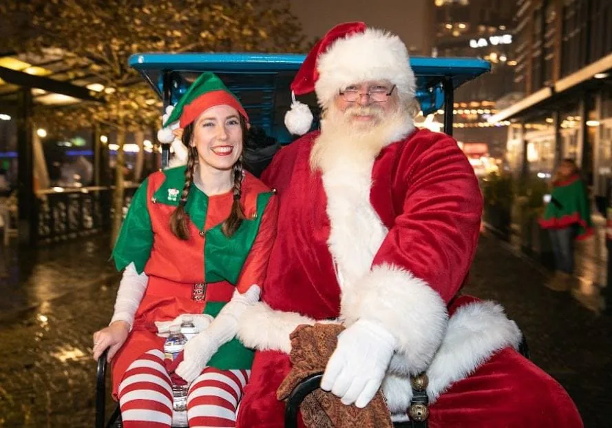 Santa Claus and a woman in an elf costume posing together on a bench at a nighttime holiday event