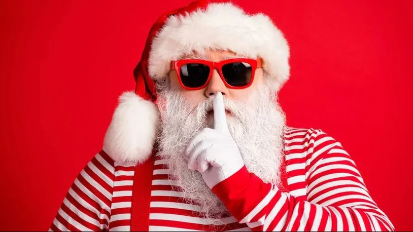 Santa in red sunglasses holding a finger to his lips in a shushing gesture on a red background