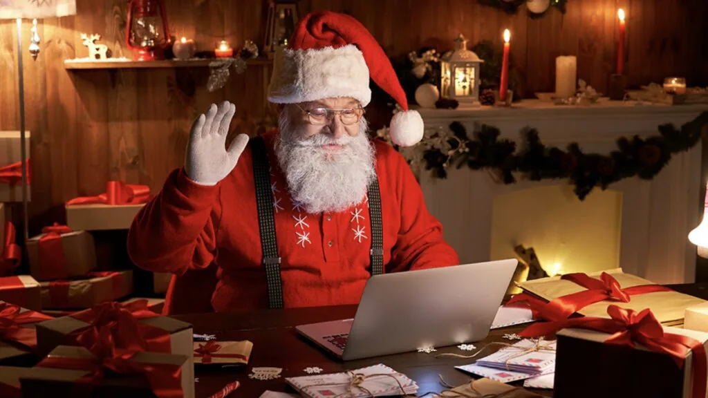 Santa Claus waving at a laptop screen at a desk surrounded by wrapped gifts and holiday decorations