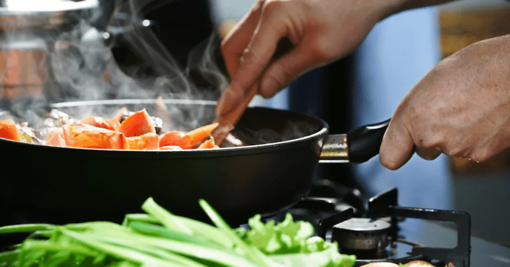 Hand stirring tomatoes in a steaming skillet on a gas stove with celery and greens in the foreground