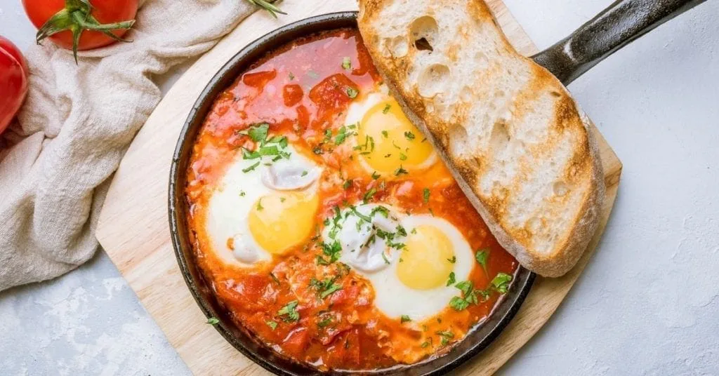 Skillet of shakshuka, eggs poached in spiced tomato sauce, with a slice of toasted bread