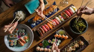 Overhead view of sushi rolls, nigiri and edamame with hands reaching in with chopsticks