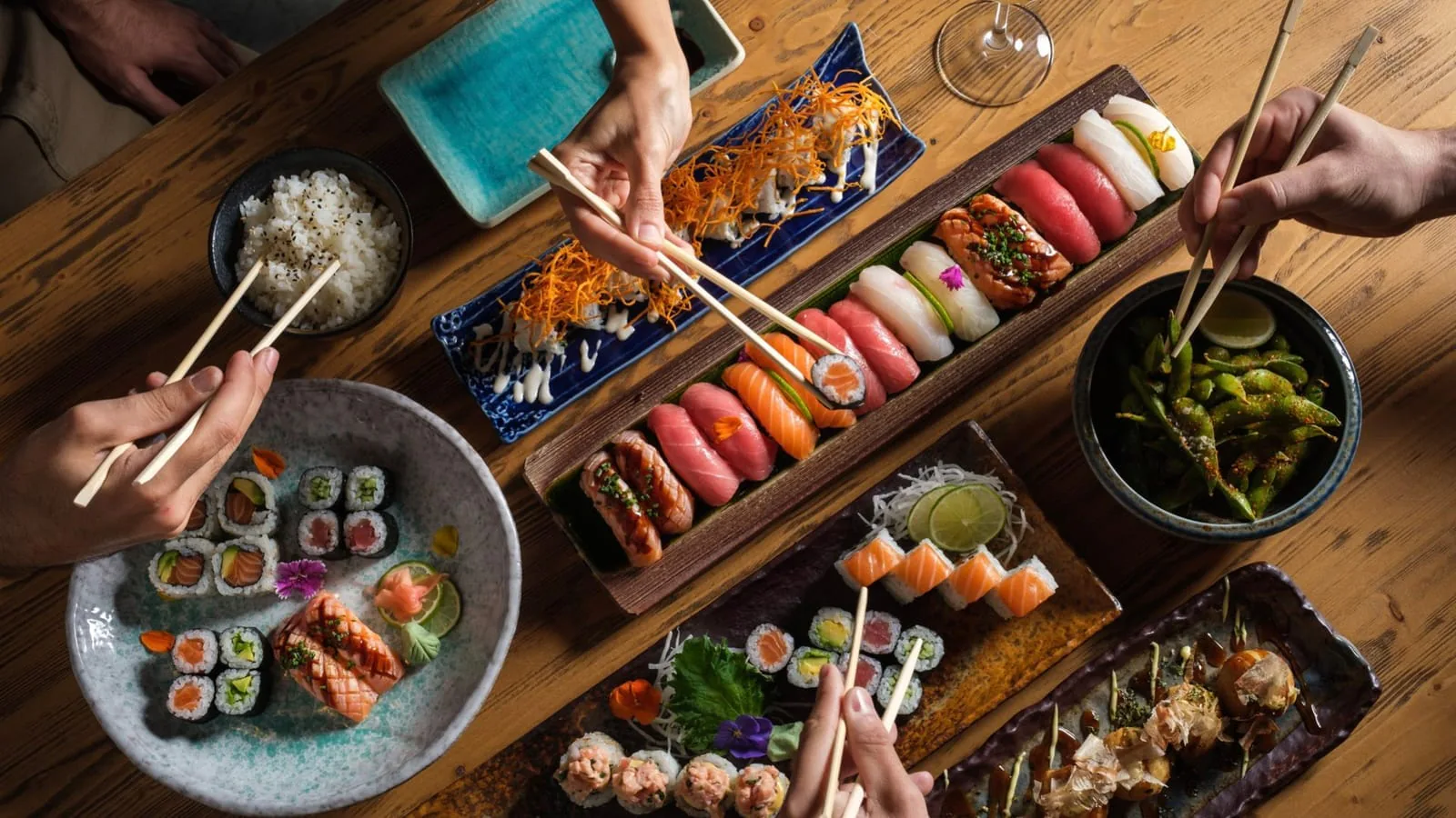 Overhead table of sushi platters, rice and edamame with several hands reaching in with chopsticks
