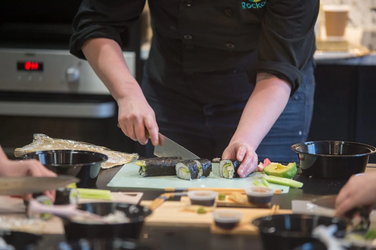 Chef in a black jacket slicing a maki roll into pieces on a cutting board at a sushi class