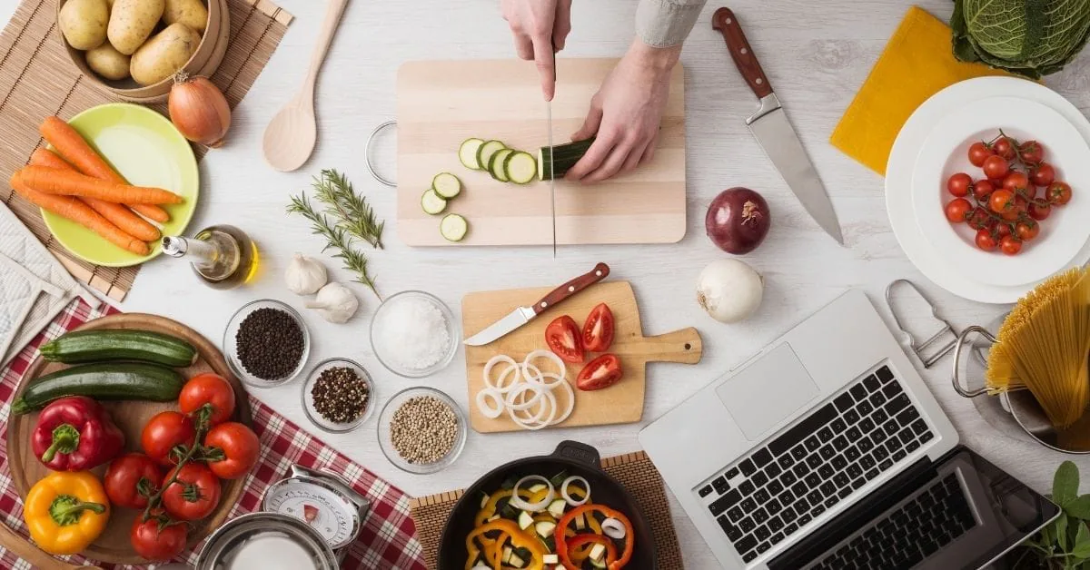 Overhead of hands slicing zucchini on a board surrounded by fresh vegetables, spices and a laptop