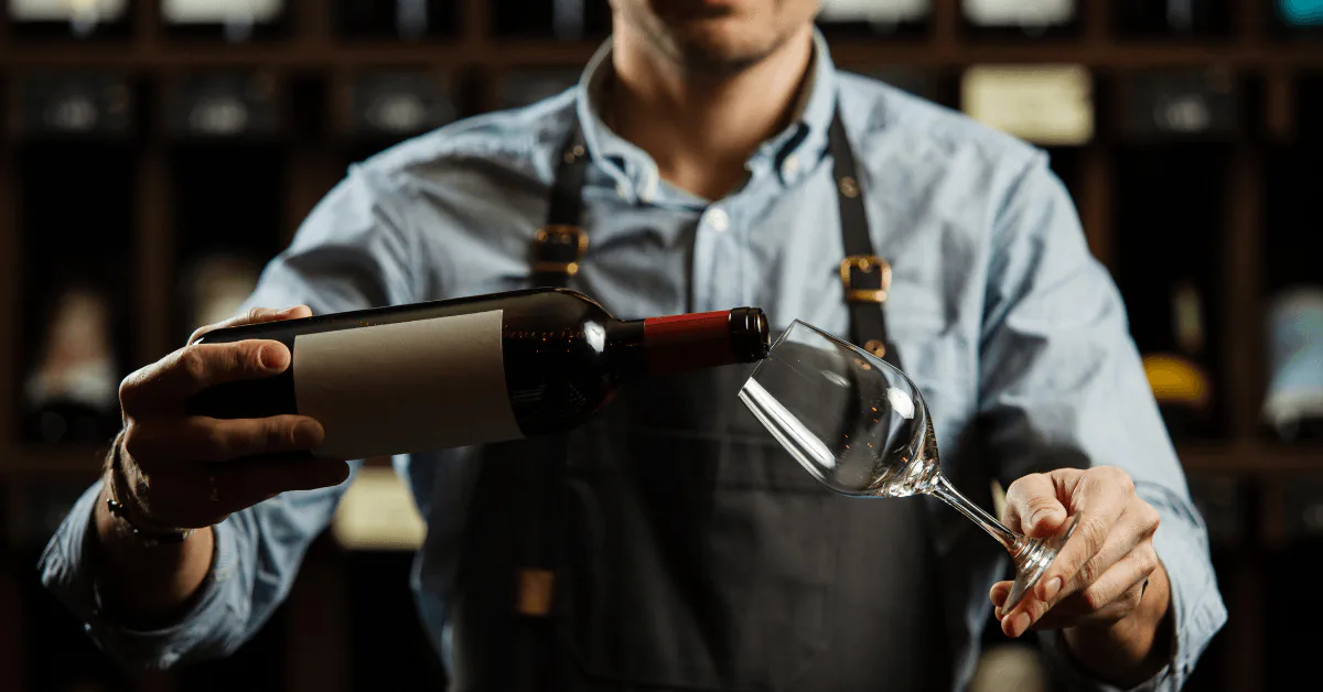 Sommelier in an apron pouring red wine into a glass in front of a wine cellar
