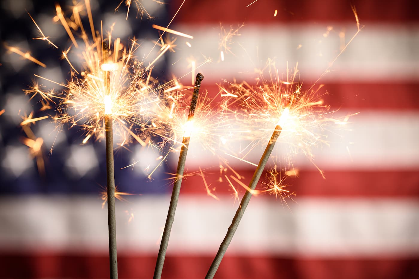 Three lit sparklers throwing sparks in front of a blurred American flag