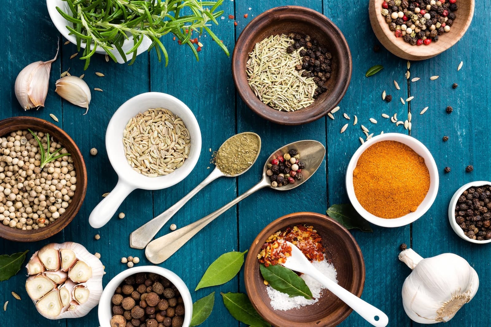 Overhead spread of spices, peppercorns, garlic and fresh herbs in bowls and spoons on a blue table