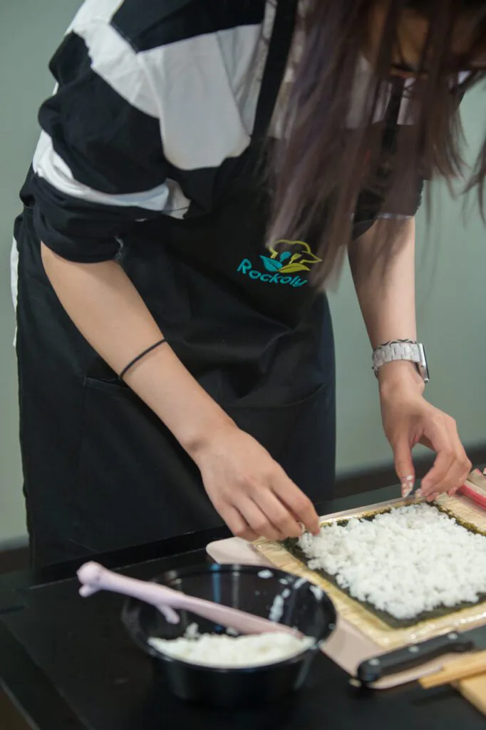 Cook in a black Rockoly apron spreading sushi rice over nori on a bamboo rolling mat