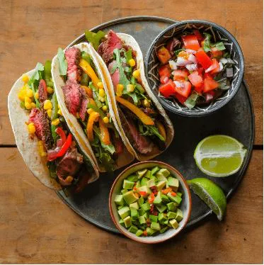 Three steak tacos on a platter with bowls of pico de gallo, diced avocado salsa and lime wedges