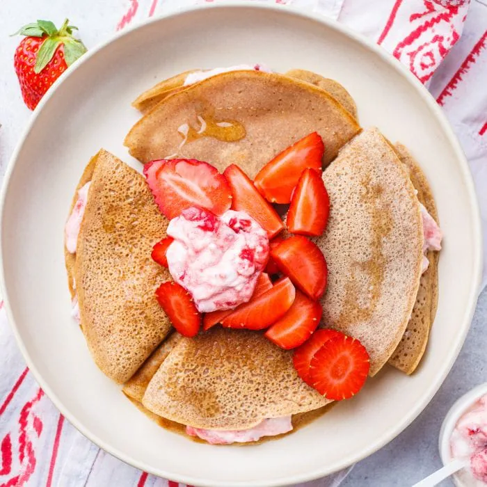 Folded crepes topped with fresh strawberries and strawberry cream on a white plate