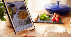 Tablet propped on a kitchen counter showing a homemade pasta recipe beside flour, basil and tomatoes