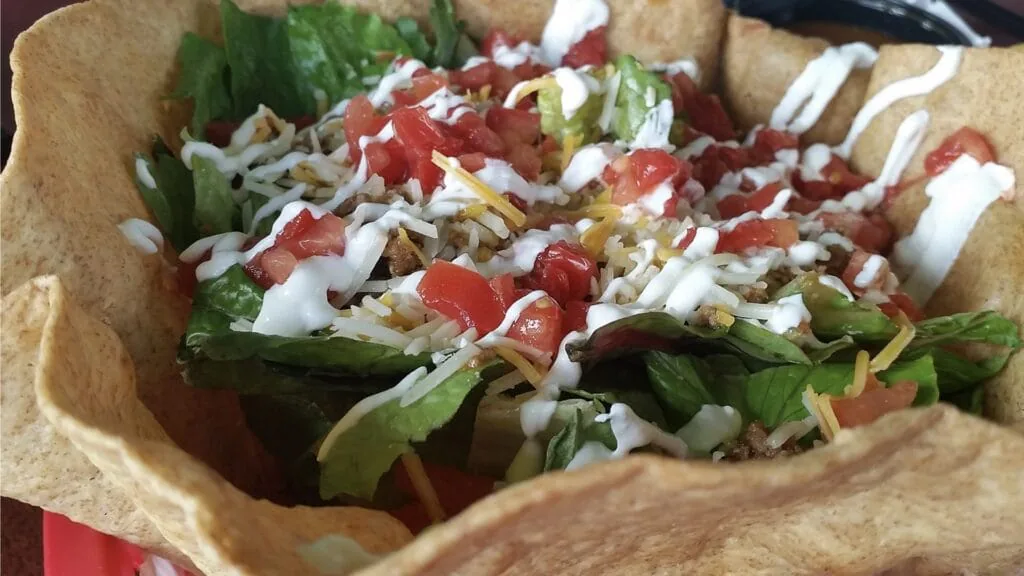 Taco salad in a crisp tortilla bowl with lettuce, diced tomato, shredded cheese and sour cream drizzle