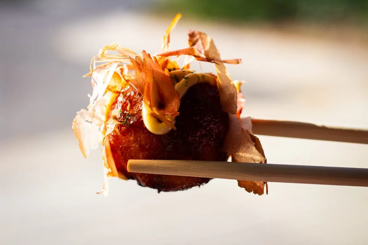 Single takoyaki ball topped with mayo and bonito flakes held up with wooden chopsticks