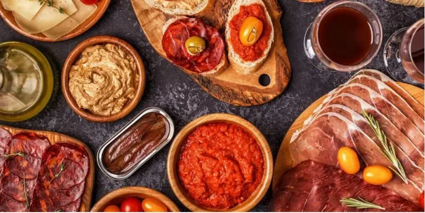 Overhead spread of cured meats, cheese, hummus, romesco dip and tomato bruschetta with a glass of red wine
