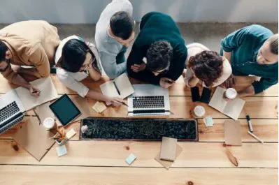Overhead view of colleagues leaning over a table with laptops, a tablet and notebooks