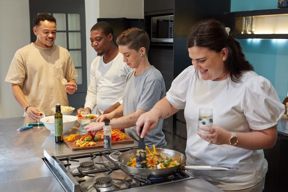Four people cooking together in a modern kitchen, one sautéing vegetables while another slices at the board