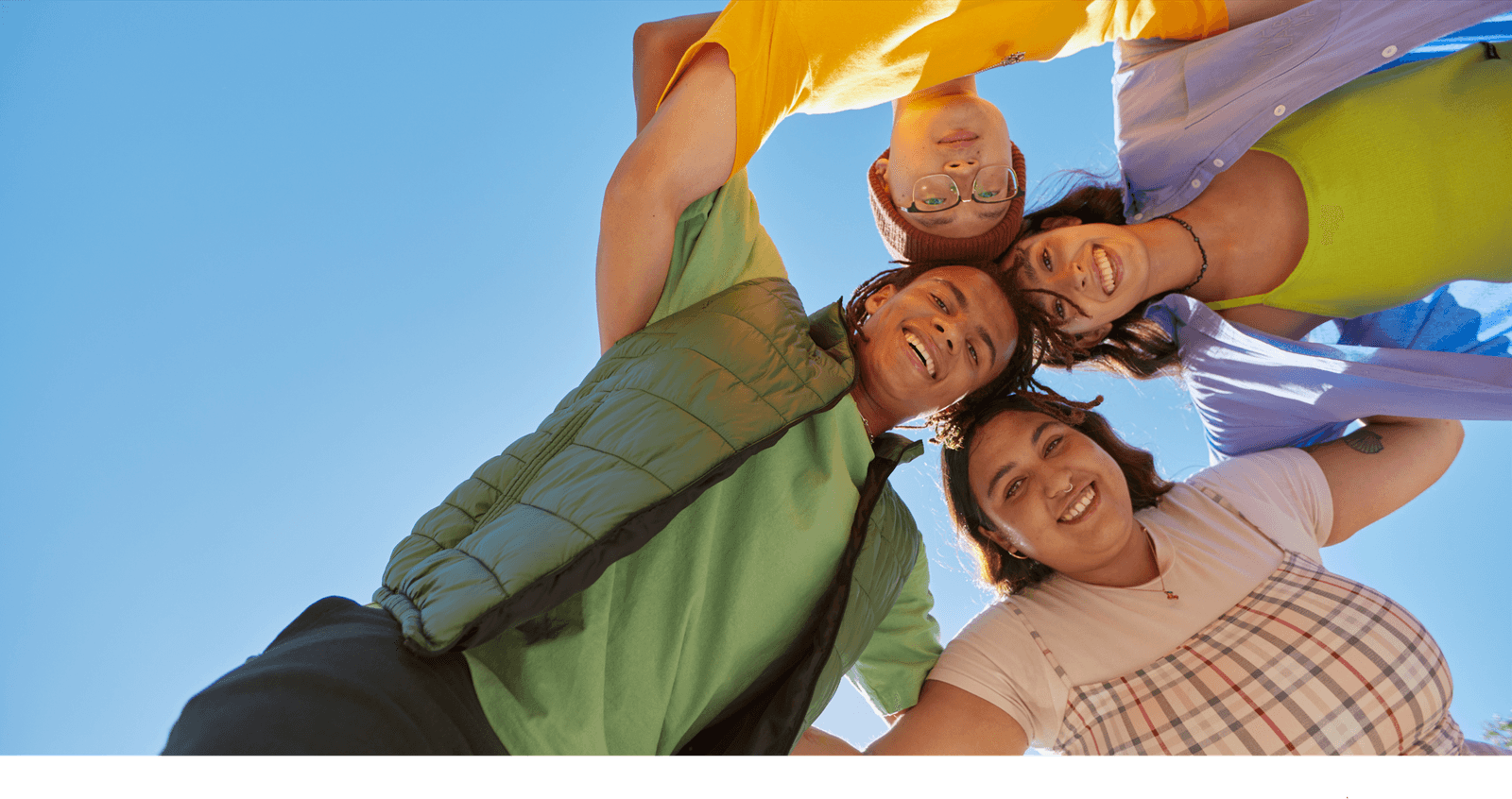 Four colleagues in a huddle circle smiling down at the camera against a bright blue sky