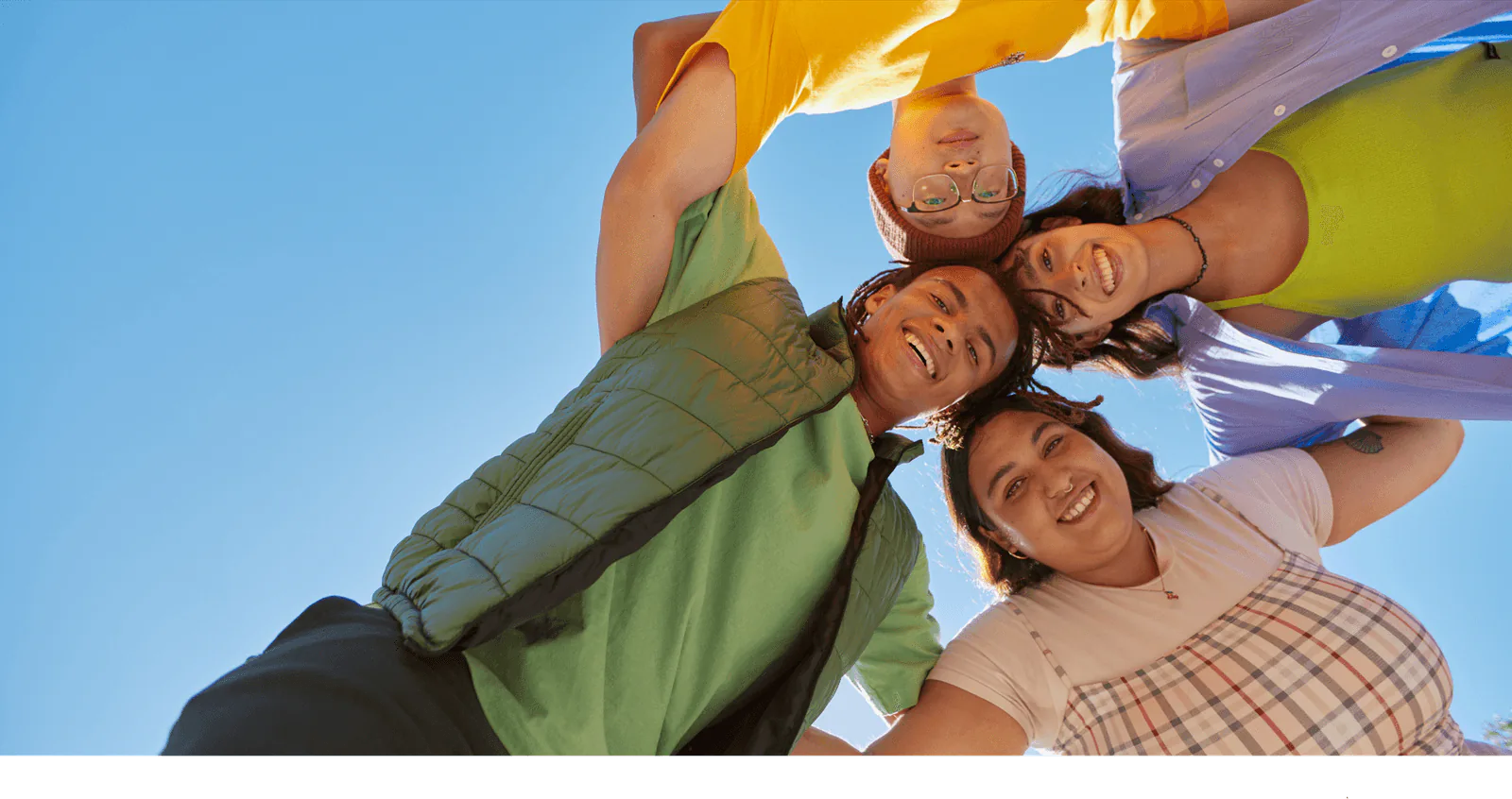 Four colleagues in a huddle circle smiling down at the camera against a bright blue sky
