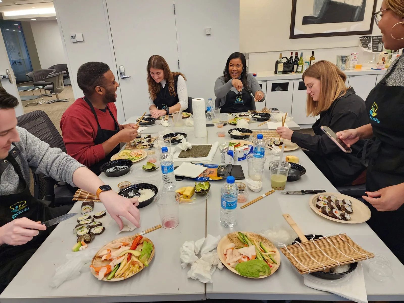 Colleagues in aprons rolling sushi at a long table with bamboo mats and plates of fish and vegetables