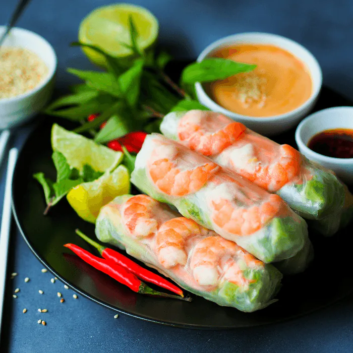 Thai fresh shrimp spring rolls on a black plate with peanut and chili dipping sauces, lime and herbs