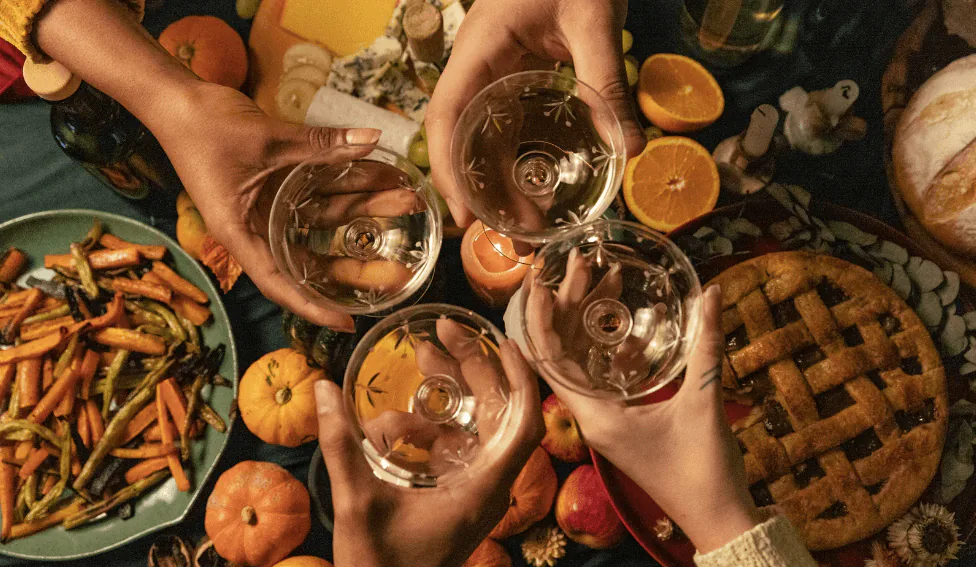 Four hands toasting wine glasses over a Thanksgiving table with a lattice pie, roasted carrots and small pumpkins