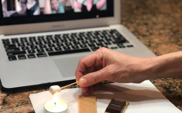 Hand toasting a marshmallow over a tea light beside a laptop showing a video call, with graham cracker and chocolate