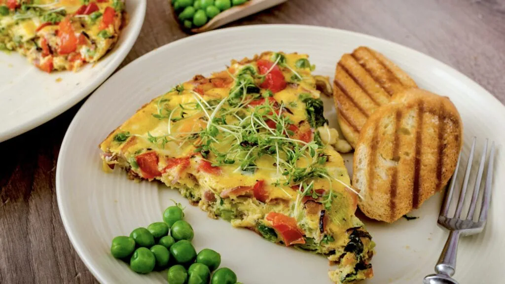 Slice of vegetable frittata topped with microgreens, served with green peas and grilled bread