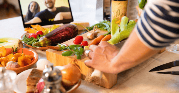 Hands prepping vegetables on a cutting board beside a laptop showing two people on a video call